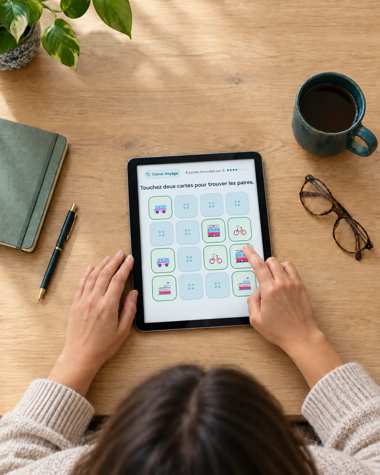 Vue de dessus : les mains d’une patiente jouent à un exercice de mémoire sur une tablette posée sur une table en bois.